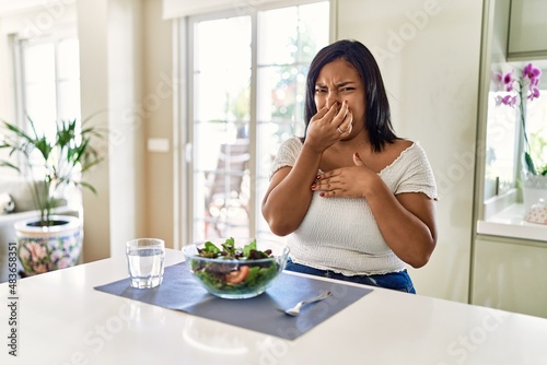 Young hispanic woman eating healthy salad at home smelling something stinky and disgusting  intolerable smell  holding breath with fingers on nose. bad smell