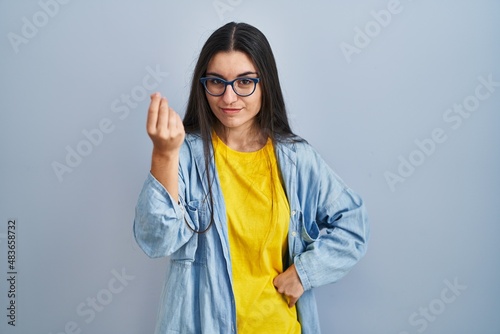 Young hispanic woman standing over blue background doing italian gesture with hand and fingers confident expression