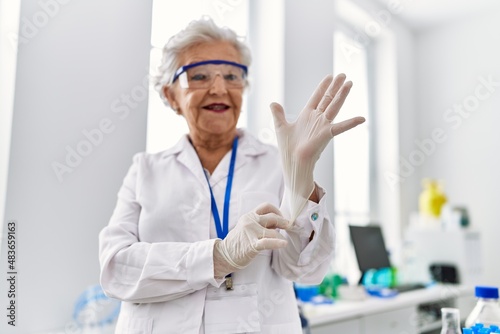 Senior grey-haired woman wearing scientist uniform wearing gloves at laboratory