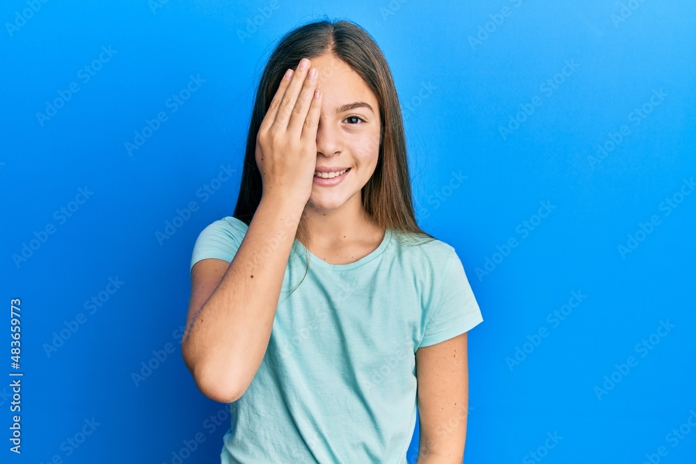 Beautiful brunette little girl wearing casual white t shirt covering one eye with hand, confident smile on face and surprise emotion. Beautiful brunette little girl wearing casual white t shirt covering one eye with hand, confident smile on face and surprise emotion.