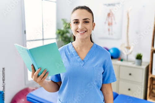 Young physiotherapist woman reading a book at pain recovery clinic looking positive and happy standing and smiling with a confident smile showing teeth