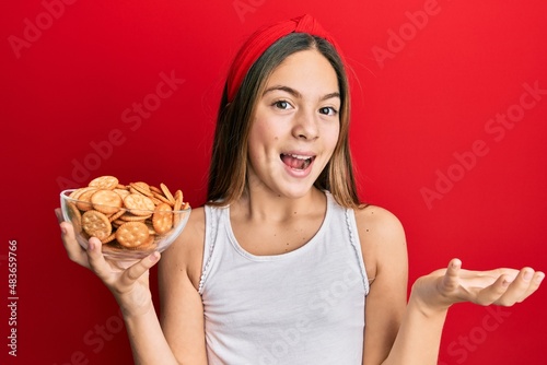 Beautiful brunette little girl holding salty biscuits in a bowl celebrating achievement with happy smile and winner expression with raised hand