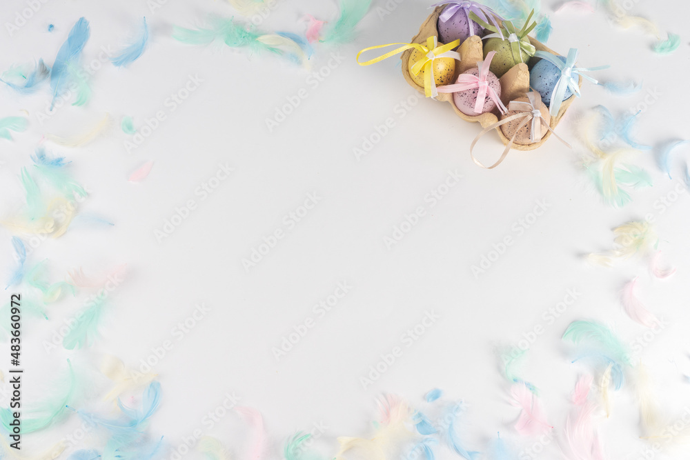 Easter colored eggs in a box, on a white background decorated with colored feathers. There is space for text. View from above.