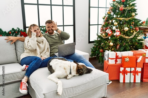 Young couple using laptop sitting by christmas tree doing ok gesture with hand smiling, eye looking through fingers with happy face. © Krakenimages.com
