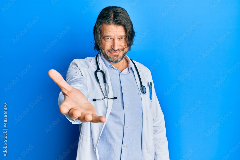 Middle age handsome man wearing doctor uniform and stethoscope smiling friendly offering handshake as greeting and welcoming. successful business. Middle age handsome man wearing doctor uniform and stethoscope smiling friendly offering handshake as greeting and welcoming. successful business.