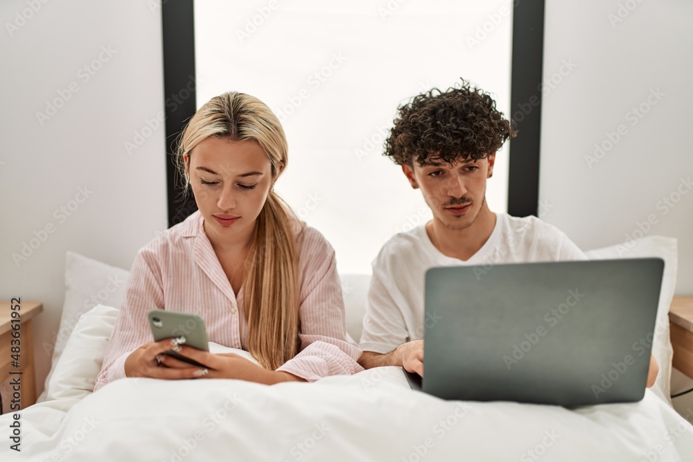Young beautiful couple using laptop and smartphone sitting on the bed at home. Young beautiful couple using laptop and smartphone sitting on the bed at home.