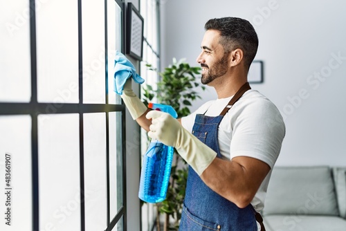 Young hispanic man cleaning window using sprayer at home