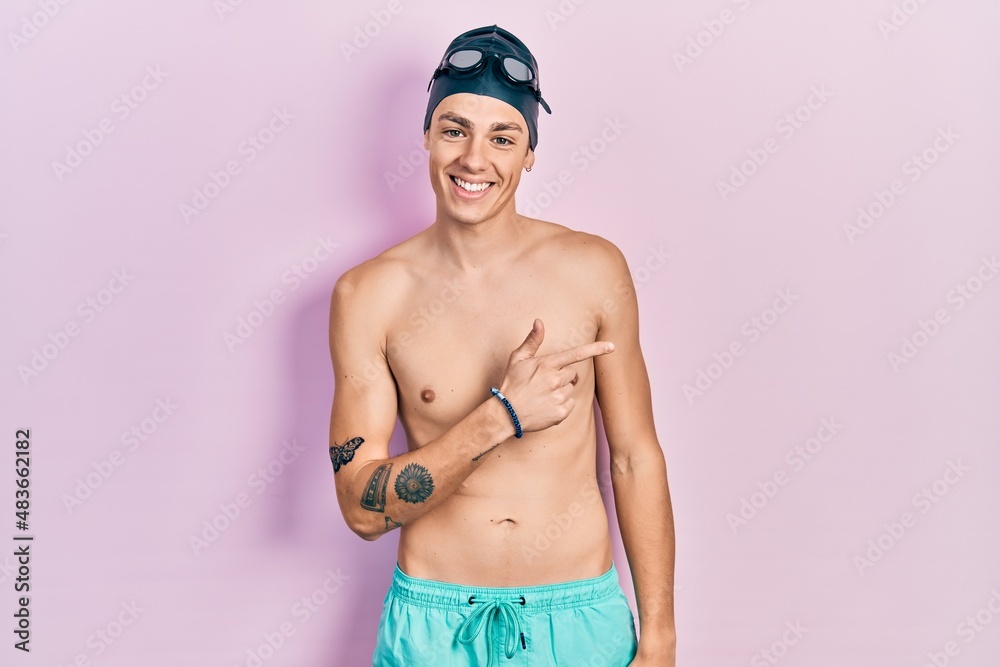 Young hispanic man wearing swimwear and swimmer glasses cheerful with a smile of face pointing with hand and finger up to the side with happy and natural expression on face