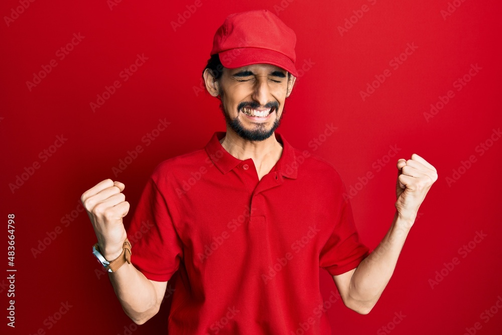 Young hispanic man wearing delivery uniform and cap very happy and excited doing winner gesture with arms raised, smiling and screaming for success. celebration concept.