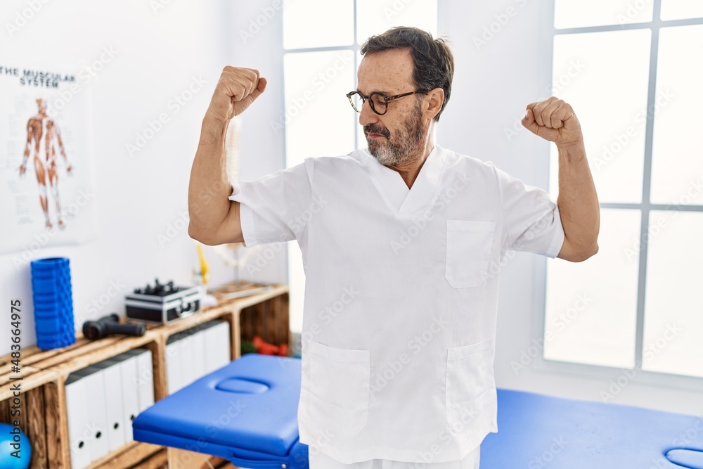 Middle age man with beard working at pain recovery clinic showing arms muscles smiling proud. fitness concept.