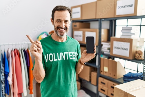 Middle age man with beard wearing volunteer t shirt holding smartphone with a big smile on face, pointing with hand finger to the side looking at the camera.