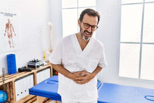 Middle age man with beard working at pain recovery clinic with hand on stomach because nausea  painful disease feeling unwell. ache concept.