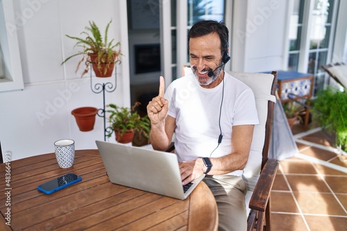 Middle age man wearing call center agent headset working from home smiling with an idea or question pointing finger with happy face, number one