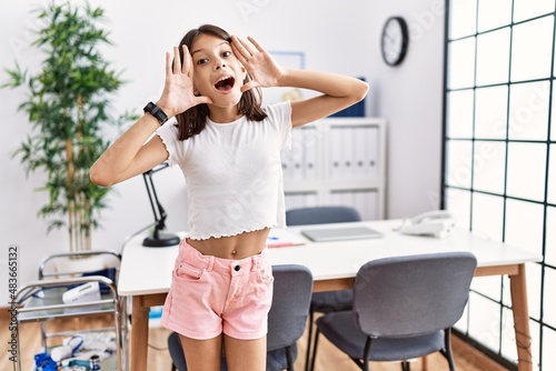 Young hispanic girl standing at pediatrician clinic smiling cheerful playing peek a boo with hands showing face. surprised and exited