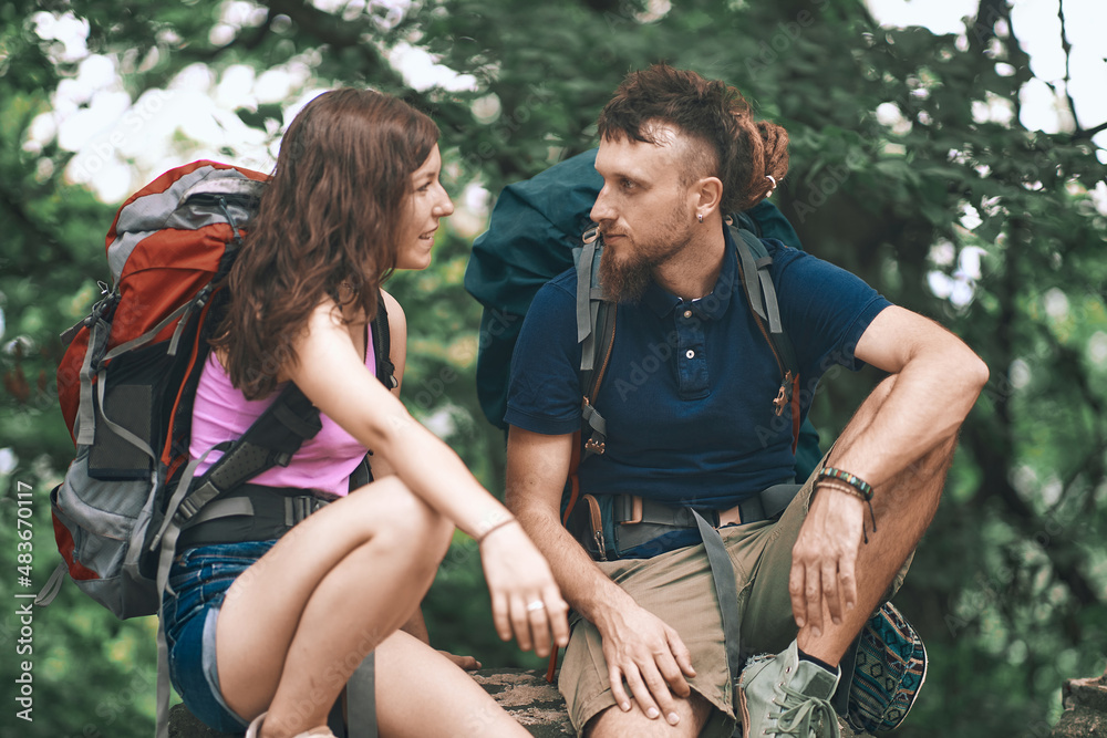 Two young friends, a man and a woman, backpacking and going on a hiking trail, looking at their cell phone, navigating through the forest