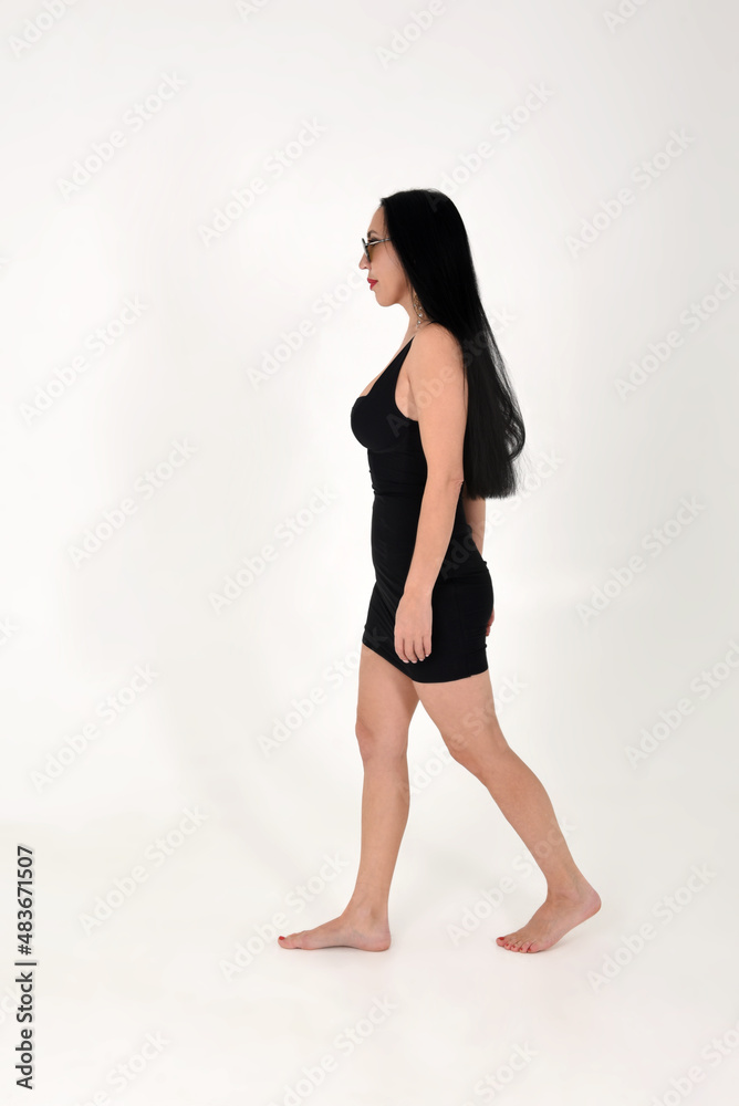 A brunette girl beautiful and stylish in a black dress is sitting walking with bare feet in the studio on a white isolated background