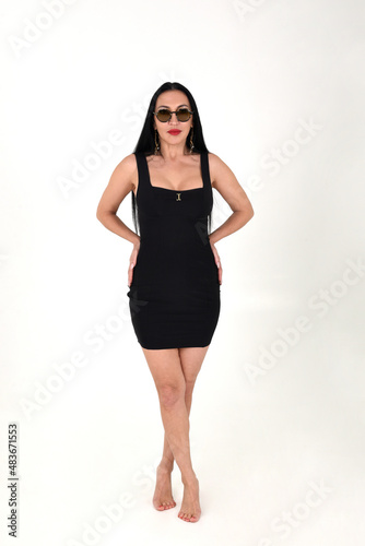 A brunette girl is beautiful and stylish in a black dress and dark glasses with bare feet in the studio on a white isolated background © Шамиль Алиев