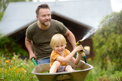 Leinwand Poster Happy little boy having fun in a wheelbarrow pushing by dad in domestic garden on warm sunny day