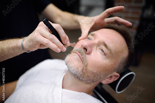 Barber master shaving handsome mature bearded man using straight razor in salon. Hair artist making beard style for person in male barbershop. Services of professional stylist.
