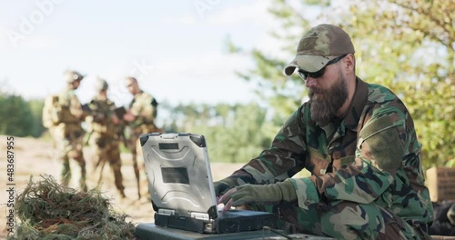Group of soldiers waiting to go into action in field dressed, prepared with equipment and weapons waiting in distance for commander, who sits at computer and sends data to the base