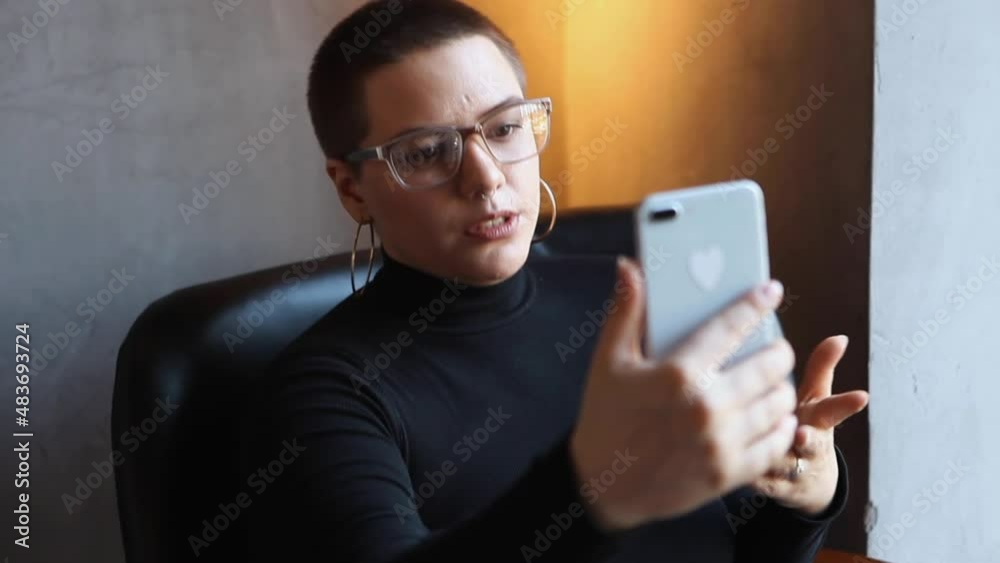 Young girl with short hair and nose piercing sits in a cafe and talking via video link on his phone. The girl leads a live broadcast or stream Young girl with short hair and nose piercing sits in a cafe and talking via video link on his phone. The girl leads a live broadcast or stream