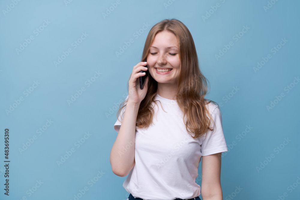 Joyful caucasian young woman talking on phone on a blue background, mobile connection. Cute smiling girl in a white t-shirt. Joyful caucasian young woman talking on phone on a blue background, mobile connection. Cute smiling girl in a white t-shirt.