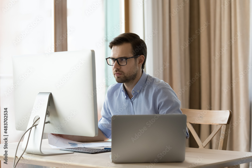 Concentrated young businessman in eyeglasses working on different devices, analyzing information, considering problem solution, creating new online project or doing economic research at home office.