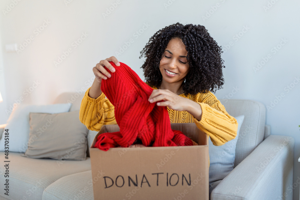 Happy African young woman sit on couch stuck clothes in donation box at