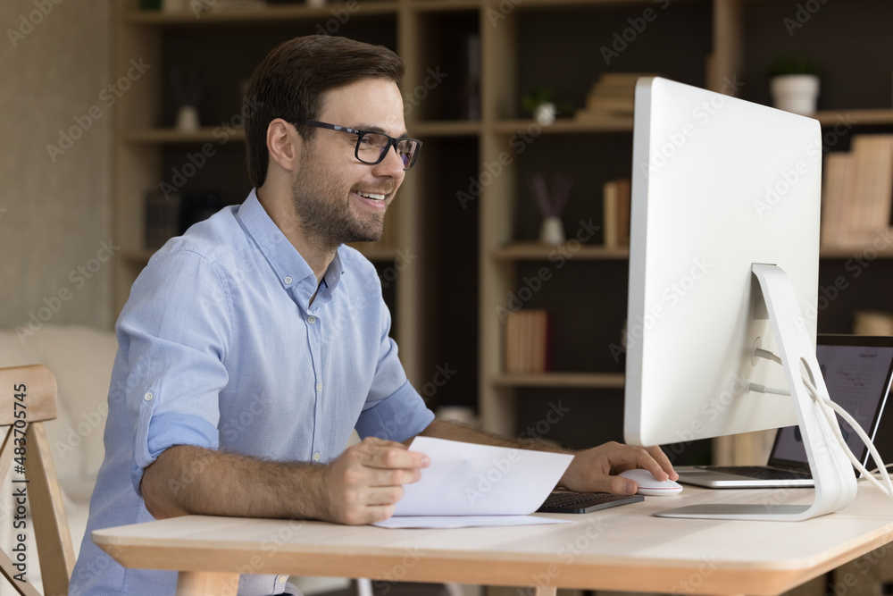 Happy young businessman in eyeglasses working on computer, analyzing paper document, feeling excited analyzing economic research report, satisfied with results or data statistics in modern home office Happy young businessman in eyeglasses working on computer, analyzing paper document, feeling excited analyzing economic research report, satisfied with results or data statistics in modern home office