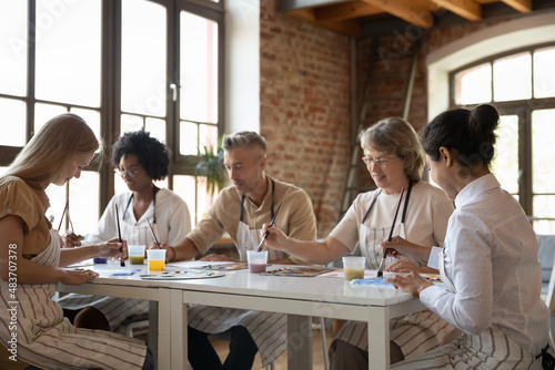 Mixed race group of different aged students drawing on art school class, painting at table, mixing colors, blending paints on palettes, enjoying artistic hobby together, talking, chatting photo