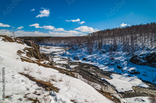 Spring landscape with river, rocks, ice, snow, trees and blue sky with white clouds