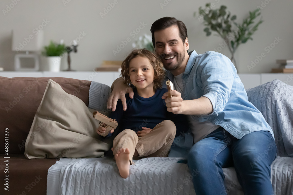 Father and son play toy guns shooting sit on sofa at home. Family spend free leisure time together, enjoy playtime. Boyish games of daddy and kid boy, pistols lovers, hobby, parent teach child concept