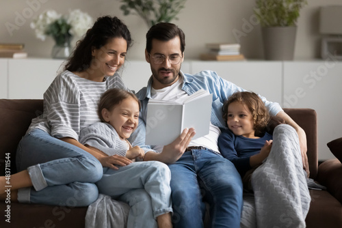 Young man his wife and cute preschool son and daughter reading book seated on sofa in living room, loving family spend leisure enjoy favourite hobby. Couple develop their kids, pastime at home concept