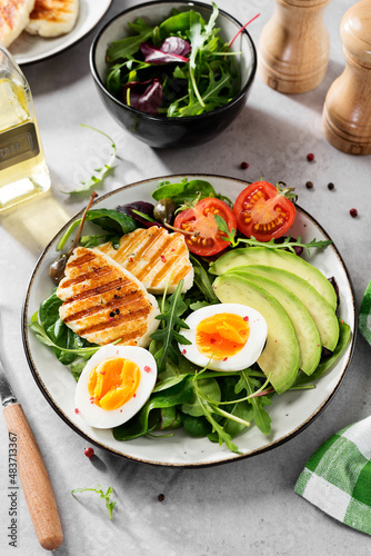 Healthy keto diet breakfast: boiled egg, avocado slices, grilled halloumi cheese, salad leaves. Light gray background.