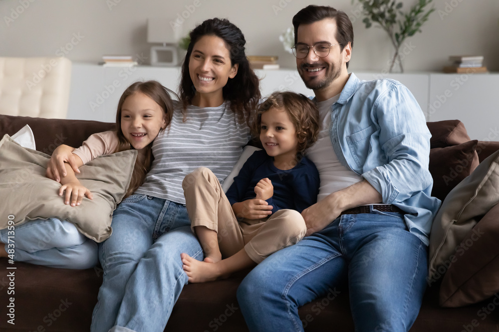 Married young couple and preschool siblings sitting on sofa relaxing together at modern home smiling looking into distance. Adoption and custody, young homeowners family portrait, bank loan concept