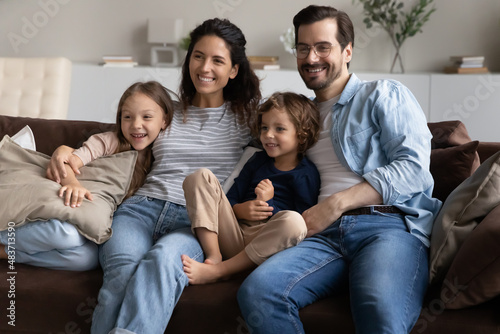 Married young couple and preschool siblings sitting on sofa relaxing together at modern home smiling looking into distance. Adoption and custody, young homeowners family portrait, bank loan concept