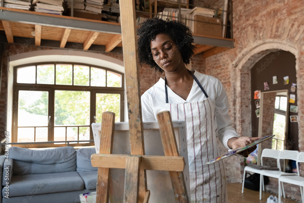Relaxed serious Afro American professional artist woman working in home craft studio, drawing at easel, holding palette, paints, creating artwork, training artistic skills. Art, talent concept