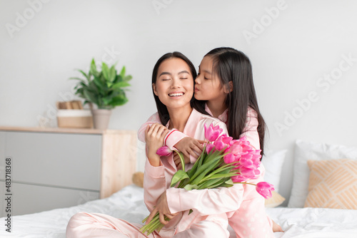Smiling happy asian teenage girl kissing and hugging millennial woman with bouquet of tulips in bedroom