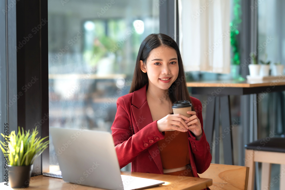 Obraz premium Confident beautiful young Asian businesswoman in a red suit sitting holding coffee with laptop computer in café. Looking at camera.