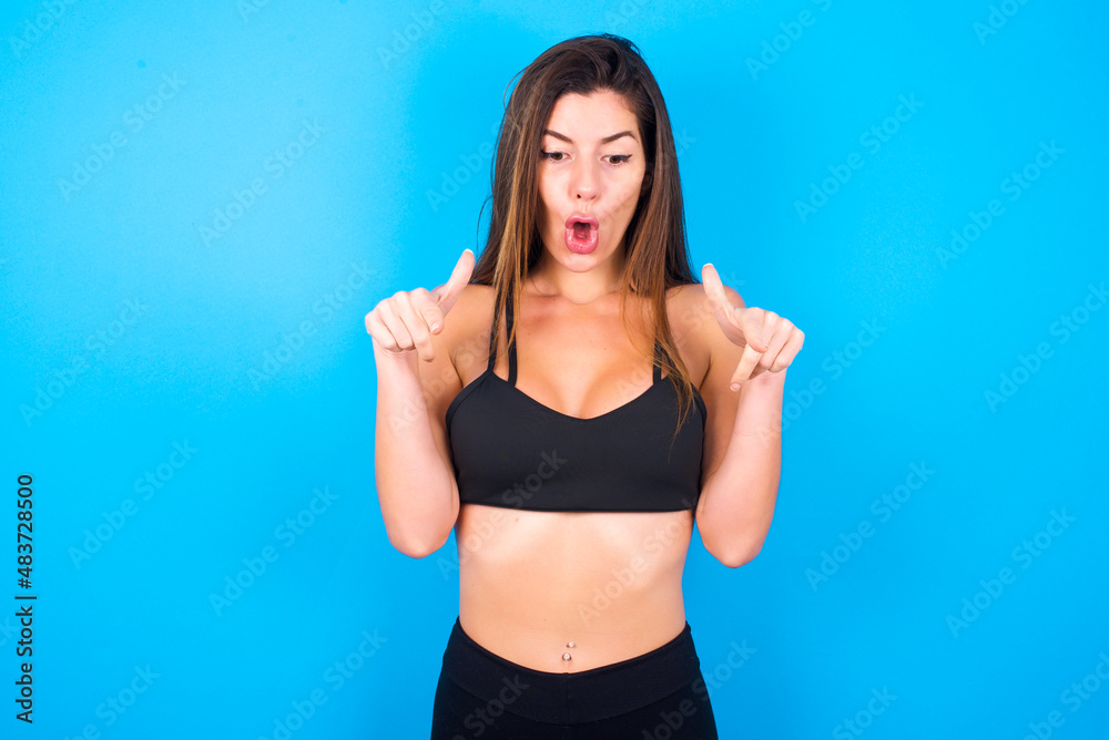 Amazed Young beautiful sportswoman doing sport wearing sportswear over blue background points down with fore fingers, opens mouth being shocked. Advertisement concept. Amazed Young beautiful sportswoman doing sport wearing sportswear over blue background points down with fore fingers, opens mouth being shocked. Advertisement concept.