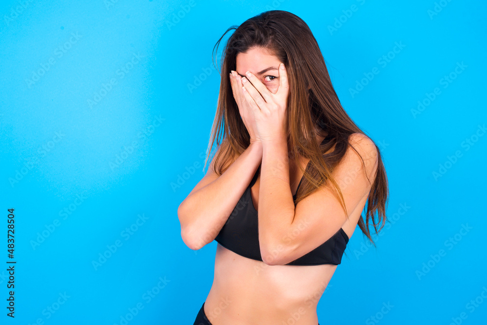 Young beautiful sportswoman doing sport wearing sportswear over blue background covering face with hands and peering out with one eye between fingers. Scared from something or someone. Young beautiful sportswoman doing sport wearing sportswear over blue background covering face with hands and peering out with one eye between fingers. Scared from something or someone.