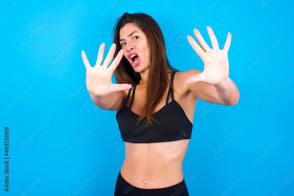 Fototapeta premium Dissatisfied Young beautiful sportswoman doing sport wearing sportswear over blue background frowns face, has disgusting expression, shows tongue, expresses non compliance, irritated with somebody.