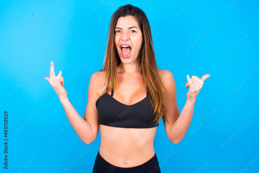 Born to rock this world. Joyful Young beautiful sportswoman doing sport wearing sportswear over blue background screaming out loud and showing with raised arms horns or rock gesture. Born to rock this world. Joyful Young beautiful sportswoman doing sport wearing sportswear over blue background screaming out loud and showing with raised arms horns or rock gesture.