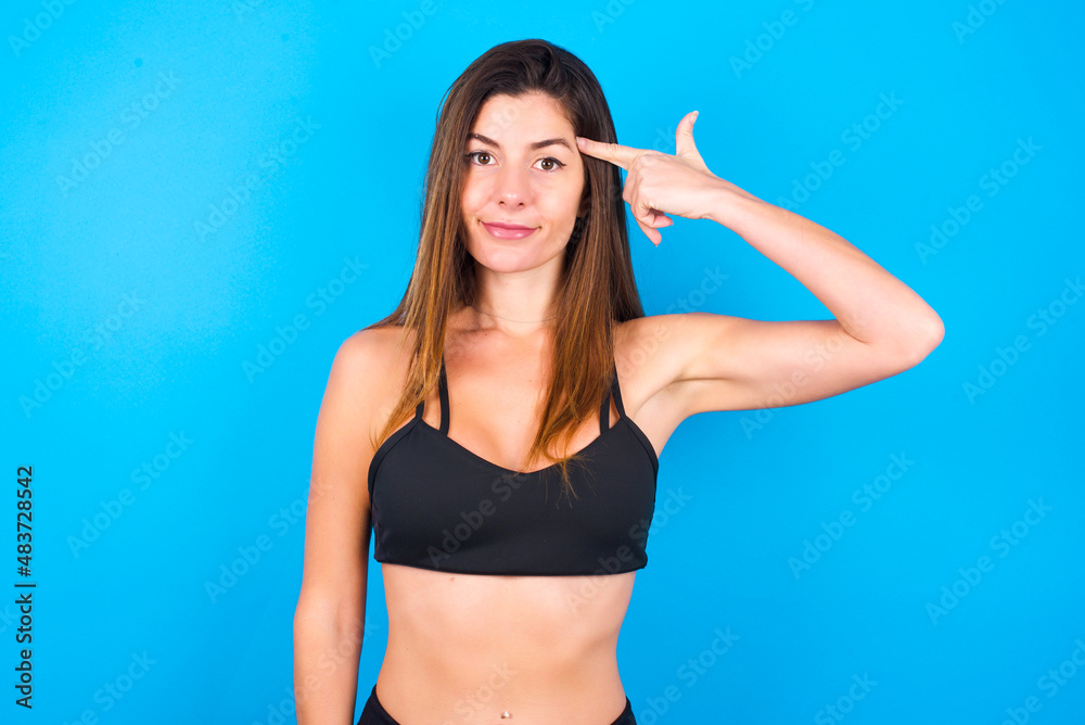 Fototapeta premium Unhappy Young beautiful sportswoman doing sport wearing sportswear over blue background curves lips and makes suicide gesture, shoots in temple with hand, tries kill himself.