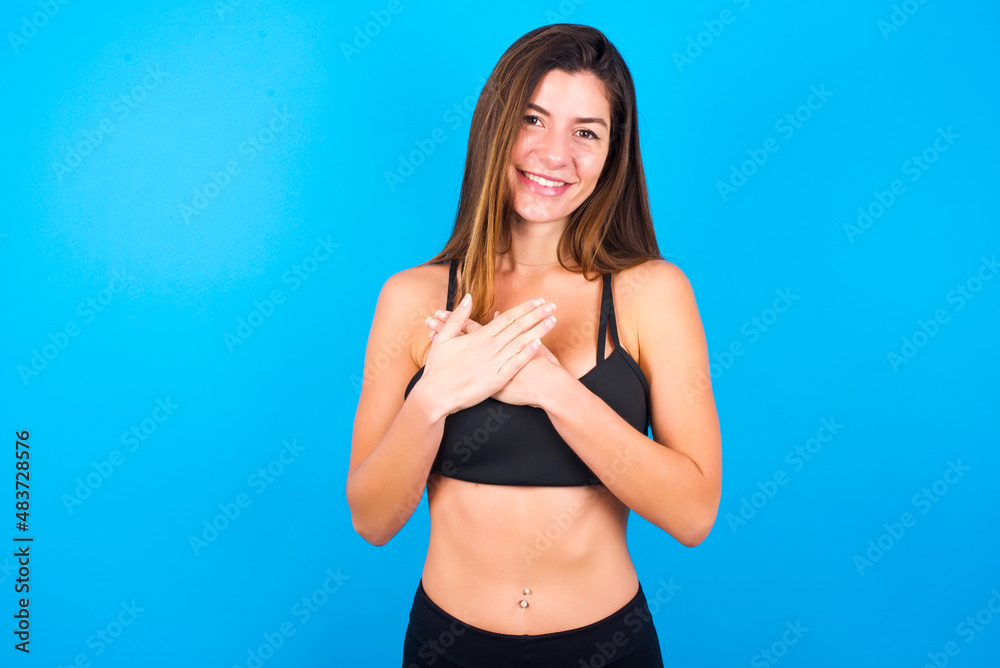 Fototapeta premium Honest Young beautiful sportswoman doing sport wearing sportswear over blue background keeps hands on chest, touched by compliment or makes promise, looks at camera with great pleasure.
