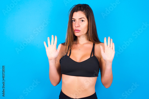 Young beautiful sportswoman doing sport wearing sportswear over blue background doing stop sing with palm of the hand. Warning expression with negative and serious gesture on the face.