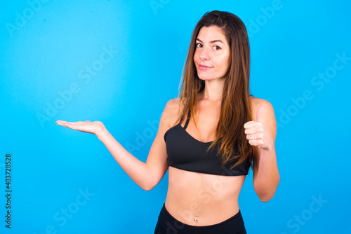 Happy cheerful Young beautiful sportswoman doing sport wearing sportswear over blue background showing thumb up and pointing with the other hand. I recommend this.