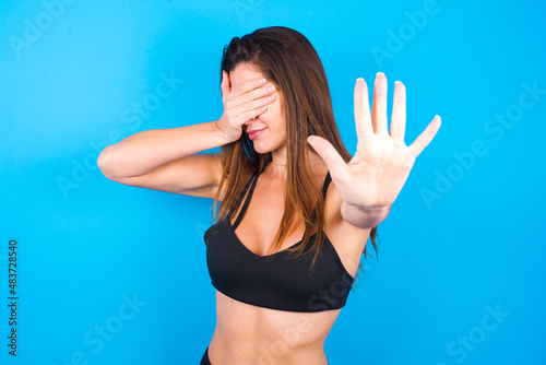 Young beautiful sportswoman doing sport wearing sportswear over blue wall covers eyes with palm and doing stop gesture, tries to hide. Don't look at me, I don't want to see, feels ashamed or scared.