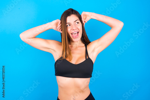 Young beautiful sportswoman doing sport wearing sportswear over blue background relaxing and stretching, arms and hands behind head and neck smiling happy