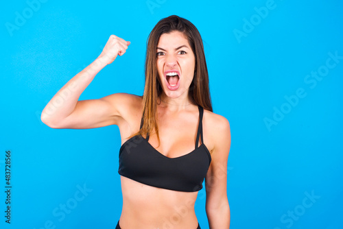 Fierce Young beautiful sportswoman doing sport wearing sportswear over blue background holding fist in front as if is ready for fight or challenge, screaming and having aggressive expression on face.